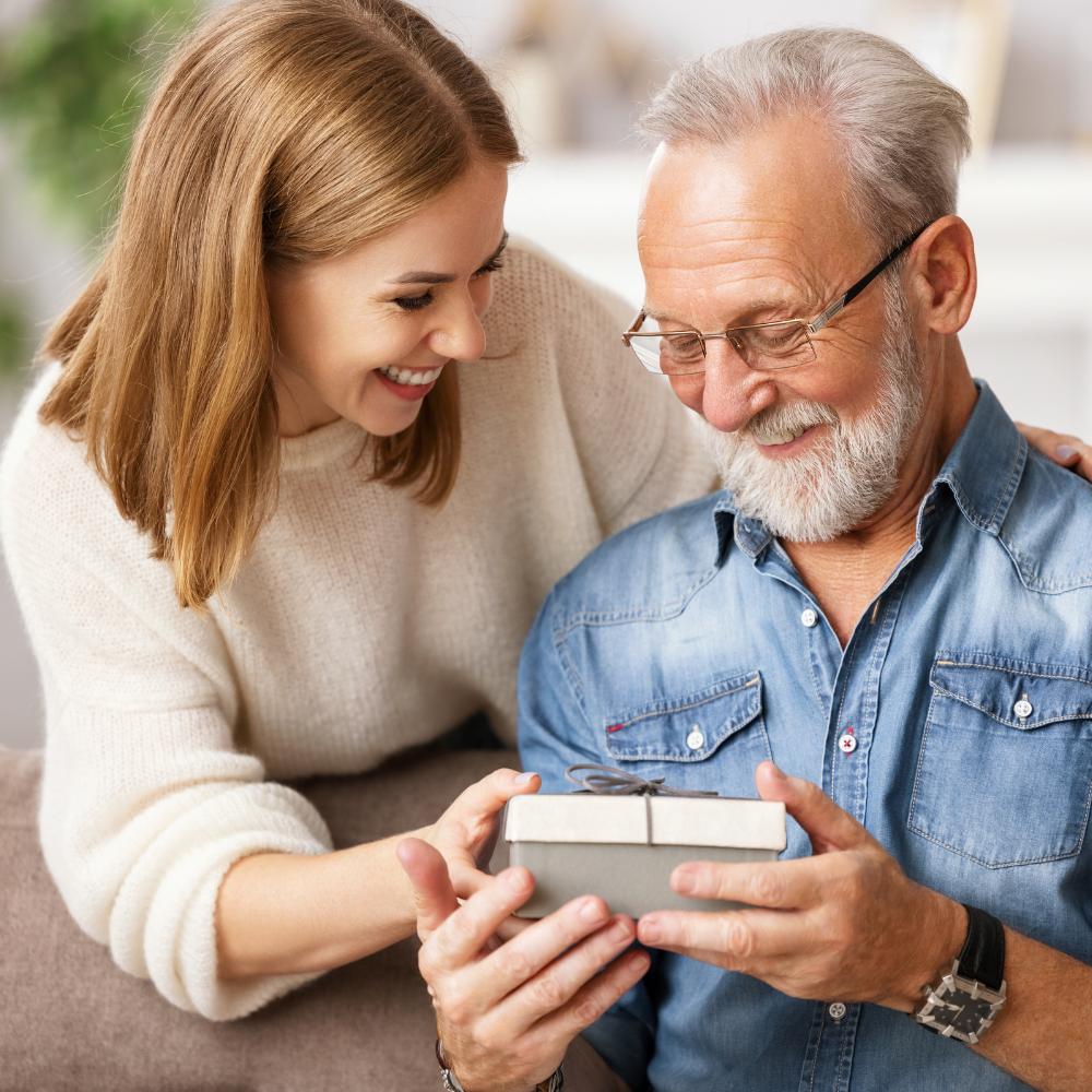 Smiling woman giving a gift box to a smiling gray headed and bearded man wearing glasses.