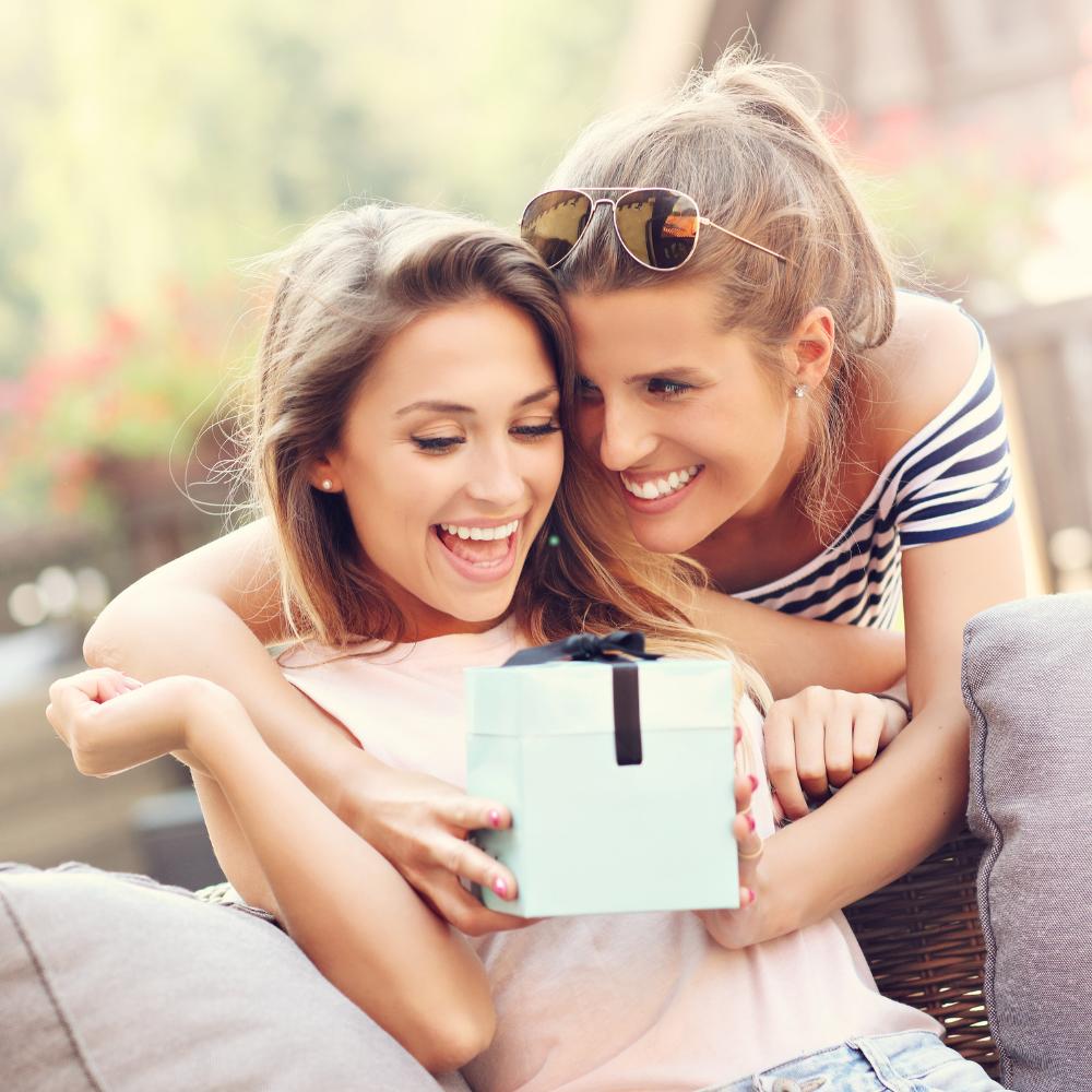 A smiling young woman puts a white gift box with a black bow in front of another young woman who is smiling in surprise.