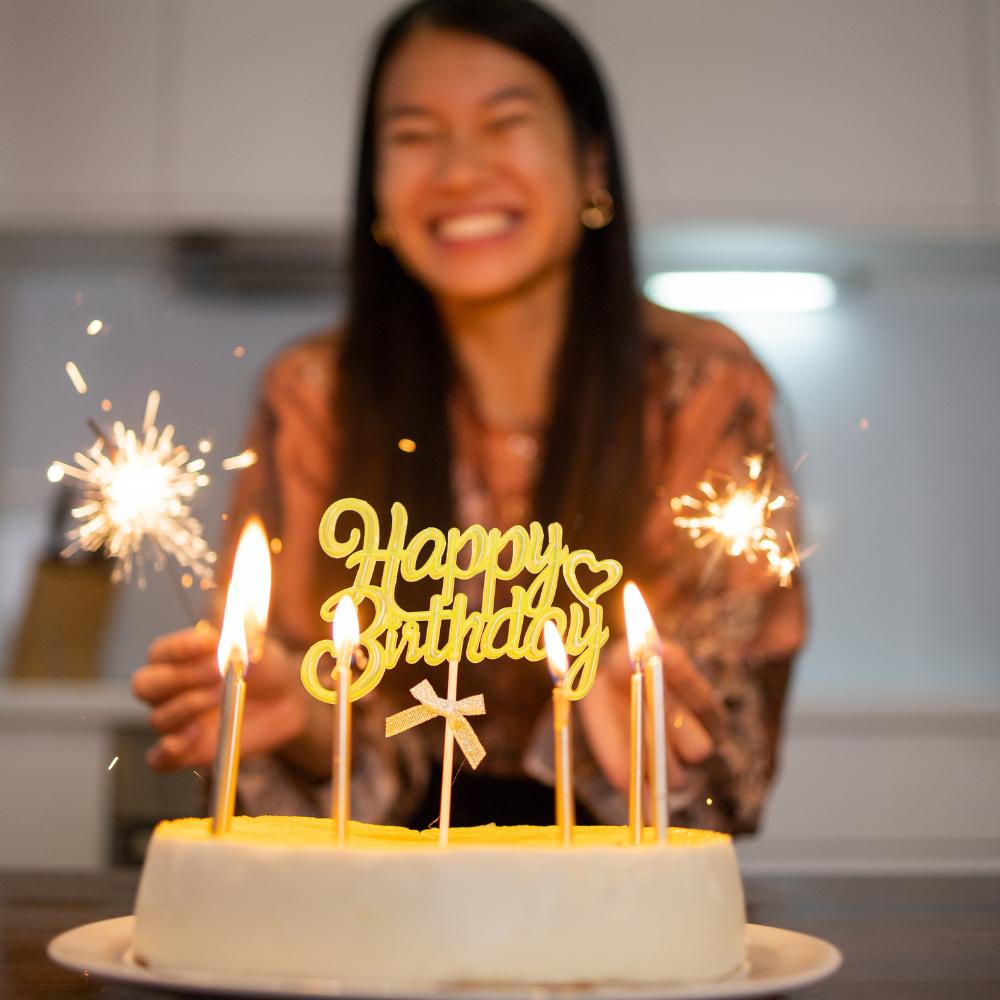 Birthday cake with lit candles and young woman in background smiling.