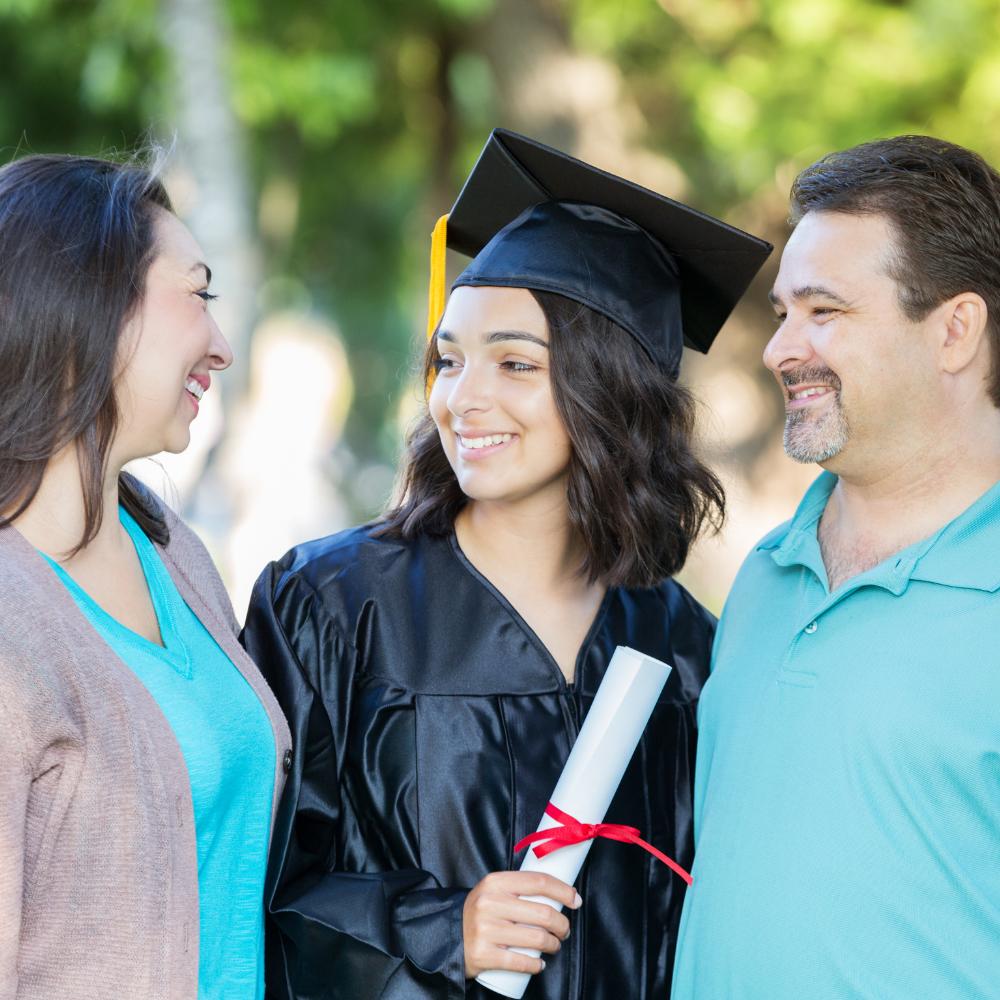 Young woman graduate wearing cap and gown standing with mother and father and everyone is smiling at each other.