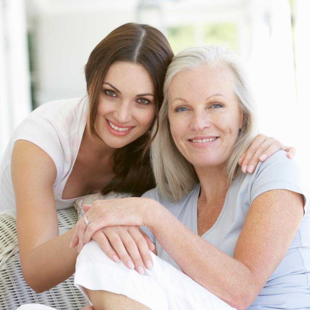 Smiling older woman sitting with smiling younger woman leaning in and hugging seated woman.