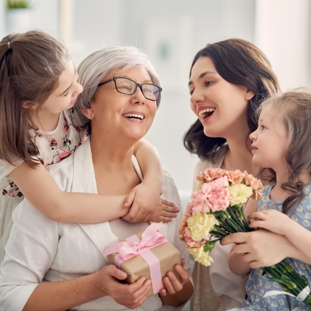 Smiling grandmother is surrounded by smiling grandchildren who are presenting her with a gift box and a bouquet of flowers.