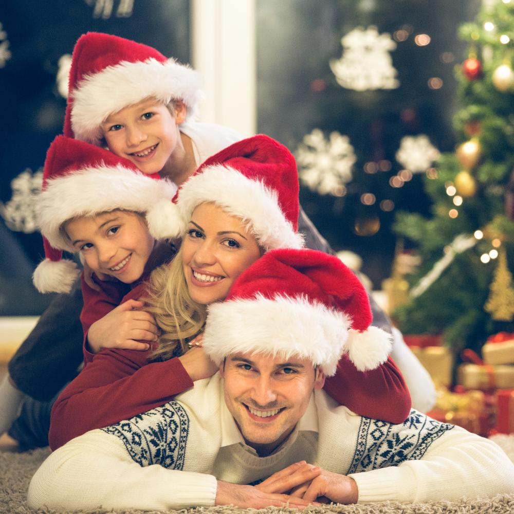 Father laying on floor in front of Christmas tree with mother resting on his back and then a daughter on her back with a son on the daughters back. All are smiling and wearing Santa hats.